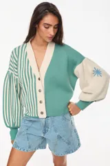 Cardigan tejido bicolor en tonos verde y crudo, con diseño de rayas verticales en la mitad derecha y liso en la izquierda. Presenta cuello en V, cierre frontal con botones y mangas abullonadas con puños ajustados.