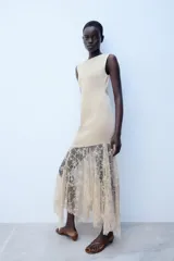 Vestido midi de color beige, con cuello redondo y diseño sin mangas. Presenta un cuerpo liso de punto con bajo asimétrico y una falda inferior confeccionada en encaje tono sobre tono.