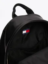 Mochila negra de poliuretano con acabado liso y diseño curvo. Tiene un compartimento principal con cierre de cremallera oculta, asa superior, correas ajustables y acolchadas, bolsillo interior de malla y bolsillo exterior con cremallera. Incluye inscripción de Tommy Jeans en las tiras e insignia de Tommy en la parte delantera.