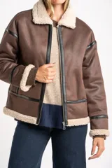 Campera tipo gamulán de corte oversize, color marrón con interior y detalles en corderito color crema. Presenta cierre frontal, cuello solapa y detalles de costuras con tiras de cuero sintético negro.