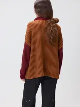 Pullover de tejido acanalado grueso con cuello alto y cierre frontal parcial. Presenta un diseño de bloques de color, con cuerpo en tono beige y mangas en contraste con rayas verticales en tonos rojizos. Incluye un detalle bordado en el pecho.