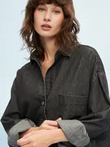 Camisa de denim azul de corte oversize, con mangas largas, cuello clásico, un bolsillo aplicado en el pecho y cierre frontal con botones metálicos.