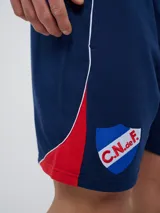 Bermuda deportiva Umbro del Club Nacional de Football, modelo Nexo para hombre, en color azul marino con detalles en rojo y blanco. Cuenta con el escudo del club bordado en la pierna derecha y el logo de Umbro en la izquierda, además de cordón ajustable en la cintura.