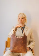 Bolso tipo matera de cuero con textura de reptil, color tostado, con manijas trenzadas y detalles de costura artesanal en los bordes.