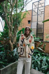 Chaqueta de algodón color crudo con cuello redondo, manga larga y bolsillos delanteros de plastrón. Presenta un diseño con bordados coloridos de flores y motivos abstractos a contraste, y cierre frontal con botones oscuros.