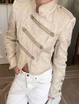 Chaqueta de estilo militar confeccionada en tejido jacquard color beige con sutil brillo. Presenta cuello mao, cierre frontal cruzado con botones decorativos, hombreras con detalles de botones y puños con botones. Posee un calce entallado y largo a la cintura.