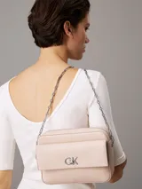 Cartera cruzada Calvin Klein de color rosa pálido, con solapa frontal y logo metálico 'CK' plateado. Posee correa de cadena plateada.