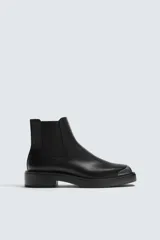 Bota tipo Chelsea de cuero negro con paneles elásticos laterales, suela de goma con volumen y detalle de pieza metálica en la puntera.