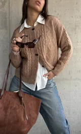 Cardigan de punto trenzado en color beige, con cuello redondo, cierre frontal de botones oscuros y mangas largas con puños acanalados.