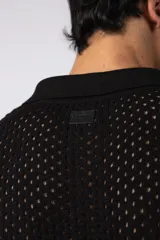 Camisa de punto calado en color beige, de manga corta y corte relajado. Presenta cuello tipo bowling, cierre frontal con botones al tono y una estructura liviana y respirable confeccionada en algodón.