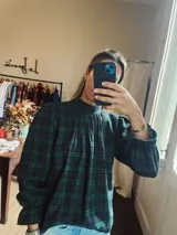 Camisa de corte holgado con estampado de cuadros en tonos verde y azul marino. Presenta cuello alto con frunces, mangas largas abullonadas con puños elásticos y diseño de tablas en el frente.