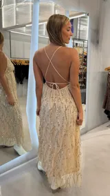 Vestido largo beige con escote en V pronunciado, tirantes finos cruzados en la espalda y diseño holgado cubierto de lentejuelas doradas dispuestas en patrones ondulados.