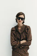 Blusa de manga larga con estampado de cuadros vichy en tonos marrón y negro. Presenta cuello redondo, cierre frontal con botones y detalle de volantes en el pecho.