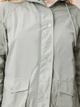 Campera de corte recto y estilo funcional, color beige, con capucha integrada, cierre frontal con botones a presión y bolsillos delanteros amplios. Presenta un diseño minimalista con cordones ajustables en la capucha.