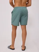Short de baño tipo volley color verde azulado, con cintura elástica y cordón de ajuste en color azul marino. Presenta un parche con el logo de la marca en la pierna izquierda y aberturas laterales en el ruedo.