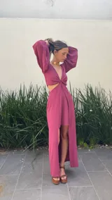 Vestido largo color bordó con escote en V profundo, detalle de nudo central y aberturas laterales en la cintura. Presenta mangas largas abullonadas con puños elásticos y falda con tajo frontal pronunciado.