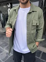 Sobre camisa verde militar con cuello clásico, cierre frontal con botones, dos bolsillos con solapa abotonada en el pecho y mangas largas con puños abotonados.