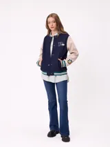 Campera estilo varsity con cuerpo en color marrón y mangas en tono beige. Presenta cierre frontal con botones a presión, cuello, puños y ruedo con franjas tejidas en verde y blanco. Incluye un parche con la letra R en el pecho y detalles bordados en las mangas.