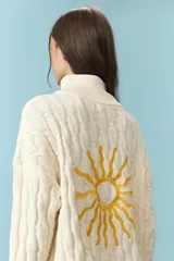 Sweater de tejido de punto con estructura de ochos en color natural, de cuello alto y manga larga. Presenta un bordado de sol en la espalda y puños con franjas de colores en rojo, celeste y amarillo.