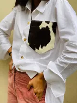 Camisa color naranja de lino o algodón con cuello clásico, cierre frontal con botones y mangas largas con puños abotonados. Presenta un bolsillo de cuero marrón en el pecho.