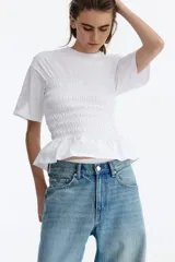 Remera de algodón blanca con hombros levemente caídos, cuello redondo y un amplio sector central con fruncido elástico que finaliza en un pequeño volante en el ruedo.