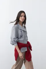 Camisa de rayas verticales con cuello mao, detalle de volados en el escote, bordados florales y recortes con calados. Posee mangas largas abullonadas con puños ajustados y cierre frontal con botones.
