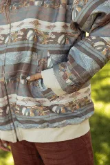 Campera de jean con cierre frontal y cuello tipo bomber, presenta un estampado horizontal con motivos paisajísticos y geométricos en tonos azules, marrones y beige. Posee puños de tela elástica en color blanco.