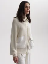 Cárdigan tejido color crema con cierre frontal de cremallera dorada, cuello alto y mangas largas raglán. Presenta dos bolsillos frontales de cuero sintético con solapa y botón a presión.