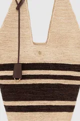 Bolso tipo hobo Anine Bing confeccionado en rafia 100% natural, con diseño a rayas en tonos beige y marrón. Cuenta con tiras de hombro integradas, detalle de tiras de cuero colgantes y un pequeño monograma metálico dorado en la parte frontal.