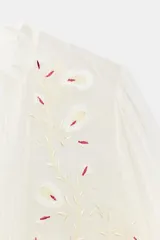 Camisa de organza de viscosa color crudo, con cuello subido, escote en V y manga larga. Presenta detalles de bordados florales en contraste a lo largo del frente y cierre frontal con botones.
