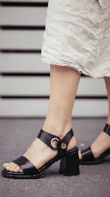 Sandalia de cuero color taupe con taco cuadrado de 6 cm, tira frontal ancha y tira en el tobillo con detalle metálico dorado y hebilla circular.