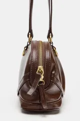 Bolso de hombro de color marrón, con diseño alargado y detalle de anillas metálicas doradas en la base de las asas. Cuenta con cierre de cremallera superior y doble asa fina para llevar colgado.