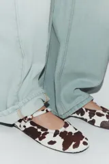 Zapato plano tipo bailarina con acabado de piel y estampado animal print de manchas marrones y blancas. Presenta una tira ajustable con hebilla metálica sobre el empeine y punta redonda.