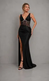Vestido largo negro de satén, con escote en V y tirantes anchos. Presenta un corpiño estilo corset translúcido con varillas visibles y detalle de pedrería brillante en el bajo busto. La falda larga tiene drapeado en la cadera y una profunda abertura lateral en la pierna.