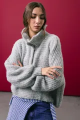 Buzo tejido en mohair de color gris, con silueta amplia y corte en línea A. Presenta cuello tipo remera con broches que permiten cerrarlo como polera, mangas largas con puños y un diseño de tejido horizontal.