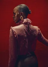 Blusa de encaje color bordó con cuello gorguera, lazo frontal y mangas largas con volantes en los puños. Presenta un diseño recto, cierre de cremallera en la parte posterior y forro interior.