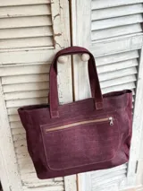 Cartera estilo shopper color bordó, confeccionada en cuero con textura tipo cocodrilo. Presenta detalles de tachas metálicas circulares y flecos laterales largos. Cuenta con doble asa de hombro.