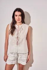 Blusa sin mangas de algodón color blanco off white, con cuello redondo y cierre frontal mediante cordones para atar. Presenta delicados detalles de bordados tonales e inserciones de encaje vertical en el frente.