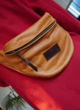 Riñonera de cuero vacuno color camel, con cierre principal y tirador de cuero, correa regulable y parche con logo grabado en el frente.
