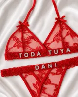 Conjunto de lencería rojo confeccionado en tul bordado, compuesto por un soutien triangular con tirantes finos para anudar al cuello, moños decorativos y aplicaciones de letras brillantes con la frase "TODA TUYA", y una bombacha colaless con la palabra "DANI".