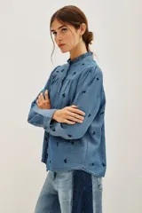 Camisa de lyocell color azul con estampado floral pequeño en tono oscuro. Presenta cuello alto con volados, cierre frontal con botones y mangas largas abullonadas con puños abotonados.