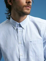 Camisa de corte clásico confeccionada en tejido oxford, con diseño de rayas verticales en tonos azul y blanco. Presenta cuello abotonado, cierre frontal con botones y bolsillo en el pecho con detalle de etiqueta bordada.