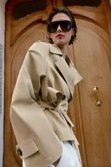 Trench corto de color beige, con cuello de solapa, manga larga con hombreras y trabillas con botones. Cuenta con cierre frontal cruzado con botones oscuros y cinturón ajustable del mismo tejido.