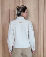 Camisa de color arena con mangas abullonadas, puños anchos con botones y cuello clásico. Presenta un diseño de corte recto con costuras visibles y cierre frontal oculto.