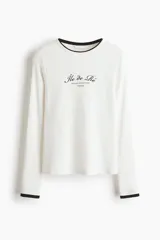 Remera de manga larga en punto acanalado fino, color blanco con ribetes negros en cuello y puños. Presenta un estampado frontal con tipografía cursiva y letras pequeñas.
