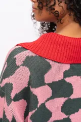 Buzo de tejido de punto con diseño estampado geométrico en tonos verde oscuro y rosado. Presenta cuello tipo polo en color rojo, cierre frontal con botones y puños acanalados en color rojo.