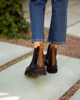 Bota tipo Chelsea de cuero negro con paneles elásticos laterales. Presenta una suela track de goma y un tirador de cuero marrón en la parte posterior.