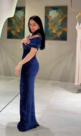 Vestido largo de fiesta en color azul, confeccionado en tela con textura arrugada. Presenta un diseño con escote tipo bardot con nudo central, hombros descubiertos y corte ajustado al cuerpo con una abertura lateral en la falda.