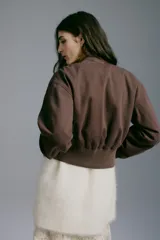 Campera bomber color marrón oscuro de tejido grueso, con cuello corto, puños y bajo acanalados. Cuenta con cierre frontal plateado, bolsillos frontales insertados y diseño de hombros caídos. Está completamente forrada.