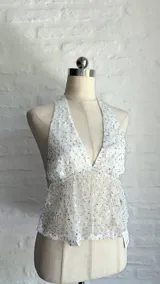 Top halter blanco con escote pronunciado en V, confeccionado en tejido transparente con lentejuelas o brillos plateados. La parte superior del busto es opaca y el resto del cuerpo es traslúcido.